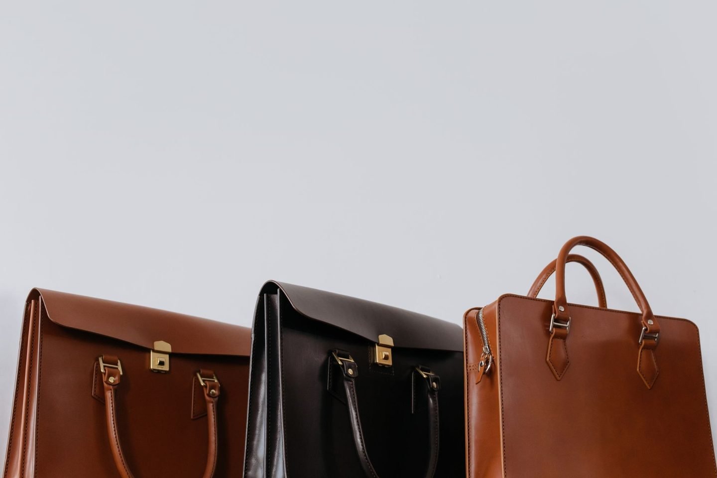 Três bolsas de couro, duas marrons e uma preta, lado a lado sobre fundo claro.
