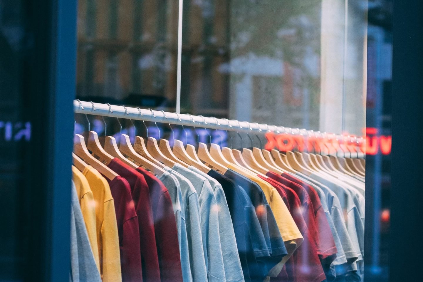 Camisetas coloridas penduradas em cabides, vistas através de uma vitrine de vidro.