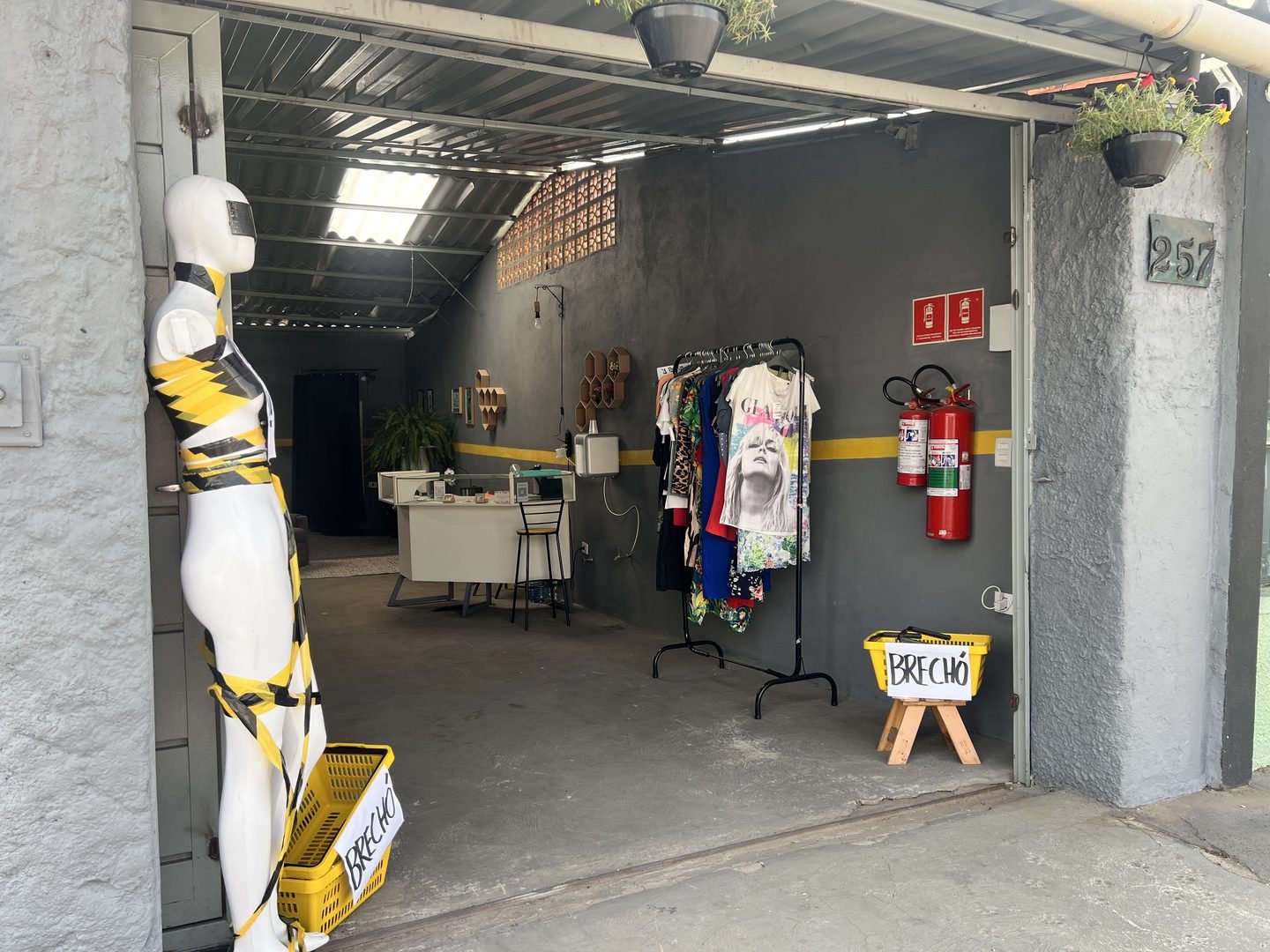 A entrada do Brechó Oficina, com o número 257 na parede, uma arara de roupas e o balcão de atendimento ao fundo
