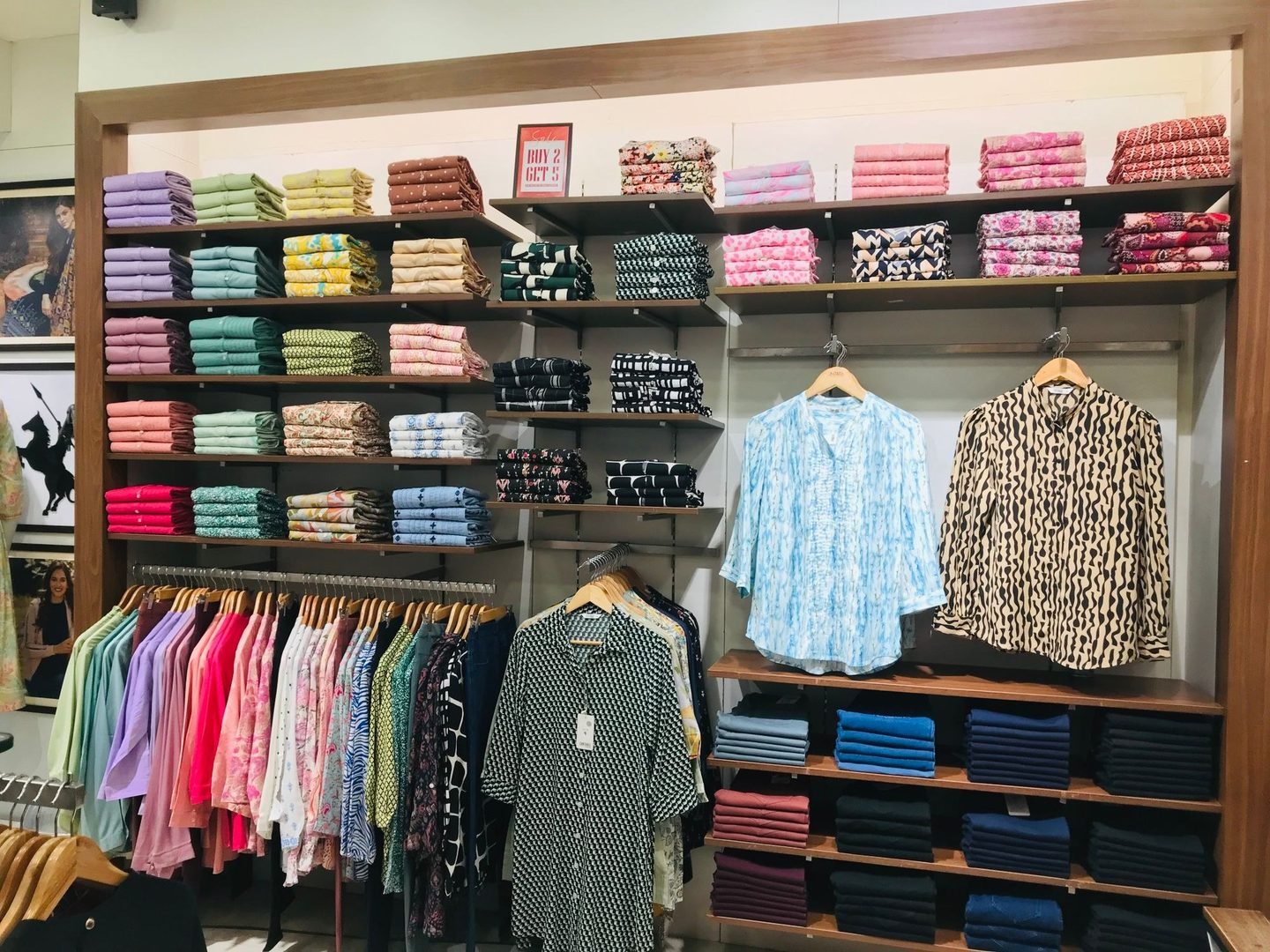 Parede de uma loja de roupas com camisas dobradas em várias cores e outras penduradas.