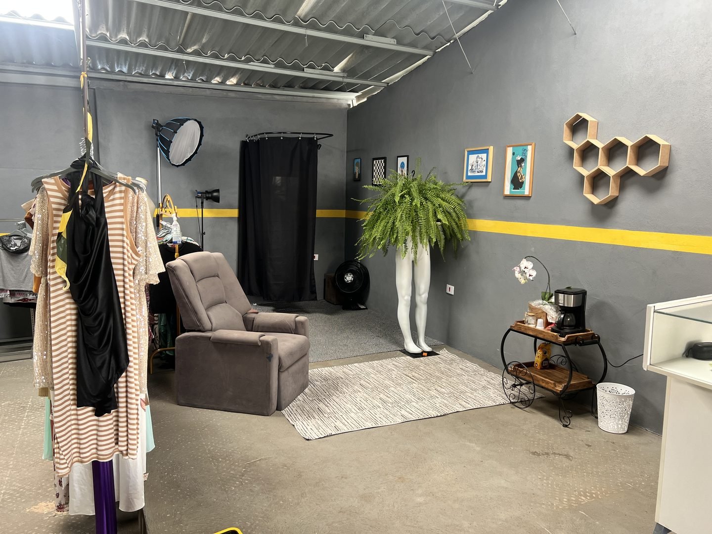 Canto do provador do Brechó Oficina, com cortina preta, poltrona, tapete e uma arara de roupas em primeiro plano