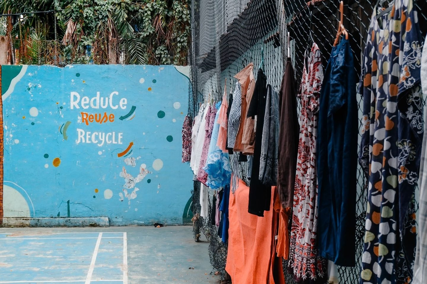 Roupas coloridas penduradas numa grade ao lado de um mural pintado com a frase Reduce, Reuse, Recycle.