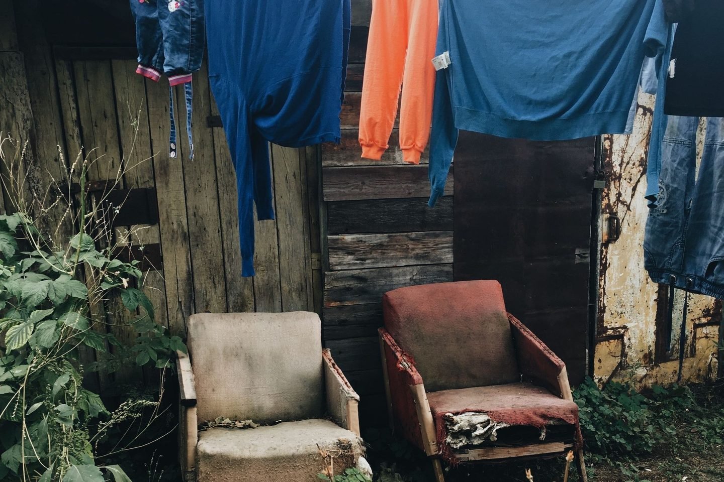 Roupas coloridas num varal diante de uma casa de madeira, com duas cadeiras velhas embaixo.