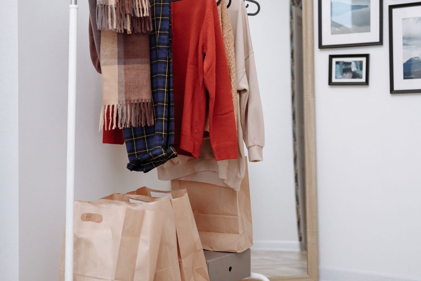 Arara branca com roupas penduradas e sacolas de papel ao lado, num ambiente claro com quadros na parede.